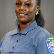 A woman in a blue public safety uniform with a badge on the shoulder, smiling, and standing against a plain background.