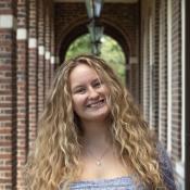 A young woman with long, curly blonde hair smiles at the camera while standing in a brick hallway with arches and hanging lanterns.