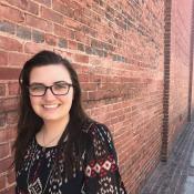 A woman with long brown hair and glasses stands smiling in front of a red brick wall on a sunny day.