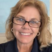 A woman with blonde hair and glasses smiles at the camera, wearing a navy collared shirt with a logo and the text "ST. MARY'S COLLEGE OF MARYLAND.