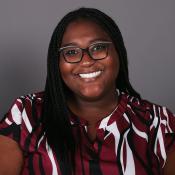 A woman with long braided hair and glasses smiles at the camera, wearing a maroon and white patterned blouse against a plain gray background.