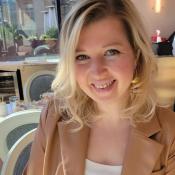 A woman with blonde hair, wearing a tan blazer and white top, smiles while sitting at a table in a sunlit café or restaurant.