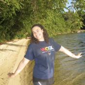 A person stands on a sandy lakeshore with arms outstretched, smiling, wearing a navy blue WGSX t-shirt, surrounded by green trees and water under a clear sky.