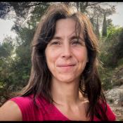 A woman with long brown hair wearing a pink shirt stands outdoors in a wooded area, smiling slightly at the camera. Trees and rocks are visible in the background.