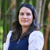 A woman with long dark hair wearing a white blouse and dark vest stands outdoors in front of blurred green foliage.
