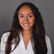 A woman with long, wavy dark hair wearing a white top is smiling in front of a plain gray background.