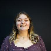 A person with long, wavy hair and a nose ring is smiling while standing in front of a plain dark background.