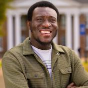 A man with short hair and a beard, wearing a green quilted jacket and smart watch, smiles with arms crossed in front of a building with white columns.