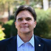 A man in a dark blazer and light blue shirt stands outside, facing the camera, with greenery and blurred buildings in the background.