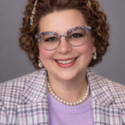 A woman with curly brown hair, wearing glasses, a beaded headband, pearl necklace, earrings, a lavender top, and a plaid jacket, smiling against a gray background.