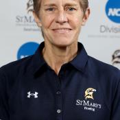 A person with short gray hair wearing a navy blue St. Mary's Rowing polo shirt stands in front of a backdrop with NCAA and St. Mary's logos.