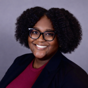 Smiling individual with glasses and curly hair wearing a dark blazer and maroon top, posing in front of a plain gray background.