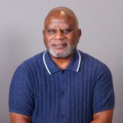 A middle-aged man with a gray beard wearing a blue short-sleeve polo shirt sits in front of a plain gray background, smiling slightly at the camera.