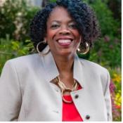 Woman with short curly hair, wearing a light beige blazer, red top, and large gold hoop earrings, smiling outdoors with greenery in the background.
