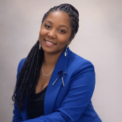 A woman with long braided hair wears a bright blue blazer, a black top, and silver jewelry, smiling at the camera.