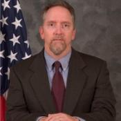 A man wearing a suit and tie sits in front of an American flag and a gray mottled background, facing the camera with a neutral expression.