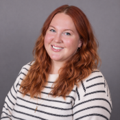 A woman with long wavy red hair, wearing a white and black striped sweater, smiling in front of a plain gray background.