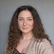 A woman with long curly brown hair, light eyes, and a nose ring, wearing a beige sweater and a necklace, poses in front of a neutral gray background.