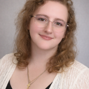 A young person with curly light brown hair, glasses, and a light lace cardigan over a black top, smiles softly at the camera against a neutral background.