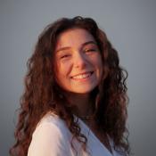 A young woman with long curly brown hair smiles at the camera, wearing a white top and a shell necklace, against a plain gray background.