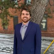 A man in a blue suit and gray tie stands outside on a snowy sidewalk in front of a brick building and a large tree.