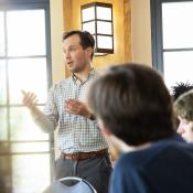 A man in a checked shirt stands and gestures while speaking to a group of seated people in a well-lit room with windows.