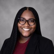 A woman with long braided hair, glasses, and a black blazer over a maroon shirt smiles at the camera against a gray background.