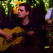 A man plays an acoustic guitar on stage with other musicians; string lights hang above them in a dimly lit venue.