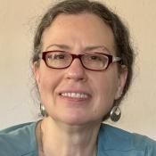 A woman with brown hair with some grey, wearing glasses, earrings and a teal top, smiling at the camera in front of a light-colored wall.