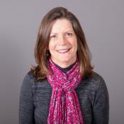 A woman with shoulder-length brown hair, wearing a gray sweater and a pink patterned scarf, smiles in front of a plain gray background.