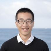 A person wearing glasses, a white collared shirt, and a black sweater smiles while standing in front of a body of water with a cloudy sky in the background.