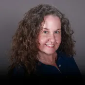 Woman with curly brown hair, blue eyes, and a blue shirt smiles at the camera against a plain gray background.