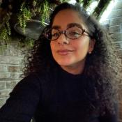 Woman with curly hair and glasses wearing a dark top, sitting indoors against a brick wall with green plants and illuminated decor in the background.