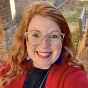 A woman with long red hair, wearing glasses, a red sweater, and blue earrings, smiles outdoors near a brick building and lawn.