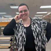 A person with short hair and glasses smiles while leaning on a file cabinet in a library, wearing a black and white patterned shirt over a black top.