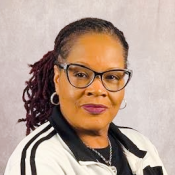 A woman with glasses and braided hair pulled back, wearing a black and white jacket, poses against a plain light background.