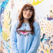 A person with long brown hair and glasses stands indoors, wearing a light blue sweater with a fish design, in front of a colorful, patterned wall.