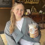 A woman wearing a gray sweater and white shirt sits at a café table, smiling and holding a large iced coffee in a clear cup. 