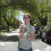 A person with glasses stands in the street holding pink flowers, surrounded by cars and leafy trees on a sunny day.