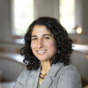 A person with curly dark hair wearing a plaid blazer and colorful necklace stands in a bright, empty classroom.