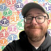 A person with a beard, glasses, and a cap smiles in front of a wall covered in colorful, repeated cartoon face designs.