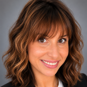 Woman with wavy brown hair wearing a black blazer and white blouse, smiling at the camera against a gray background.