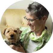 A person with short gray hair and glasses smiles while holding a small, brown dog on a beige chair.