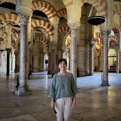 A person stands inside a historic building with striped arches, marble columns, and patterned walls, illuminated by natural light.