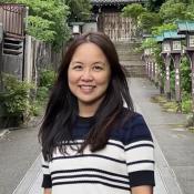 Woman with long dark hair stands smiling on a paved path lined with greenery and lanterns, with traditional wooden architecture in the background.