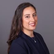 A smiling woman with long dark hair wearing a navy blue blazer and a pendant necklace.