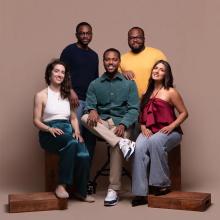 Five people pose together against a beige background, with two seated in front and three standing behind them, all smiling at the camera.