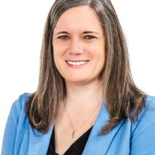 Woman with long brown and gray hair wearing a light blue blazer and black top, smiling, posed in front of a white background.