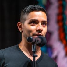 A person with short dark hair and earrings speaks into a microphone, wearing a black shirt. The colorful, blurred background suggests a lively setting.
