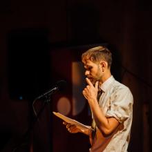 Joe Hall stands at a microphone, holding papers and touching his face thoughtfully, under warm stage lighting.